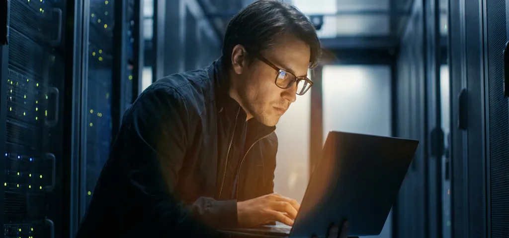 Sicherheitsexperte analysiert Laptop-Bildschirm in dunklem Serverraum mit blinkenden Rack-Servern im Hintergrund.